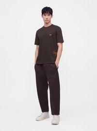 Νεαρός άνδρας με μαύρο t-shirt και μαύρο παντελόνι και άσπρα αθλητικά παπούτσια στέκεται μπροστά σε απλό λευκό φόντο.