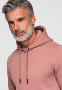 Mann mit grauem Haar und Bart, der einen staubigen rosa Hoodie trägt, leicht nach links blickend vor einem schlichten hellen Hintergrund.