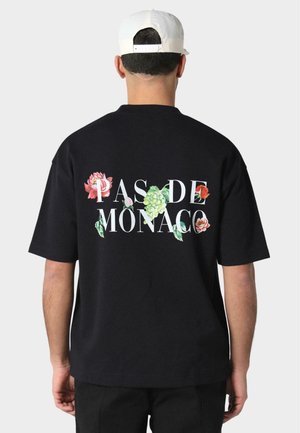 Sort bomulds t-shirt med et blomsterdesign og "PAS DE MONACO" trykt i hvidt på ryggen. Korte ærmer og afslappet pasform.