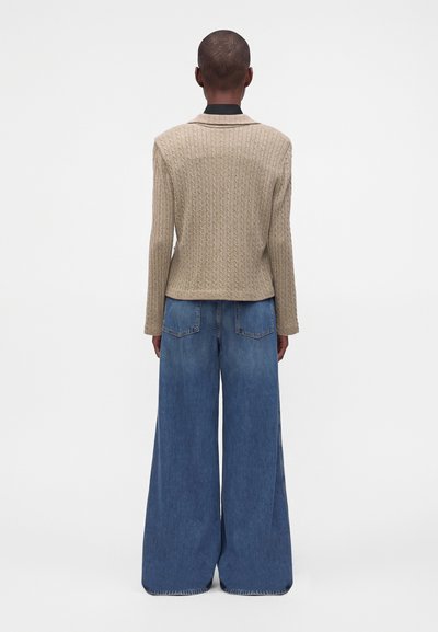 Personne aux cheveux courts portant un pull beige en maille torsadée et un jean denim bleu à jambes larges, debout, dos tourné, sur un fond blanc.