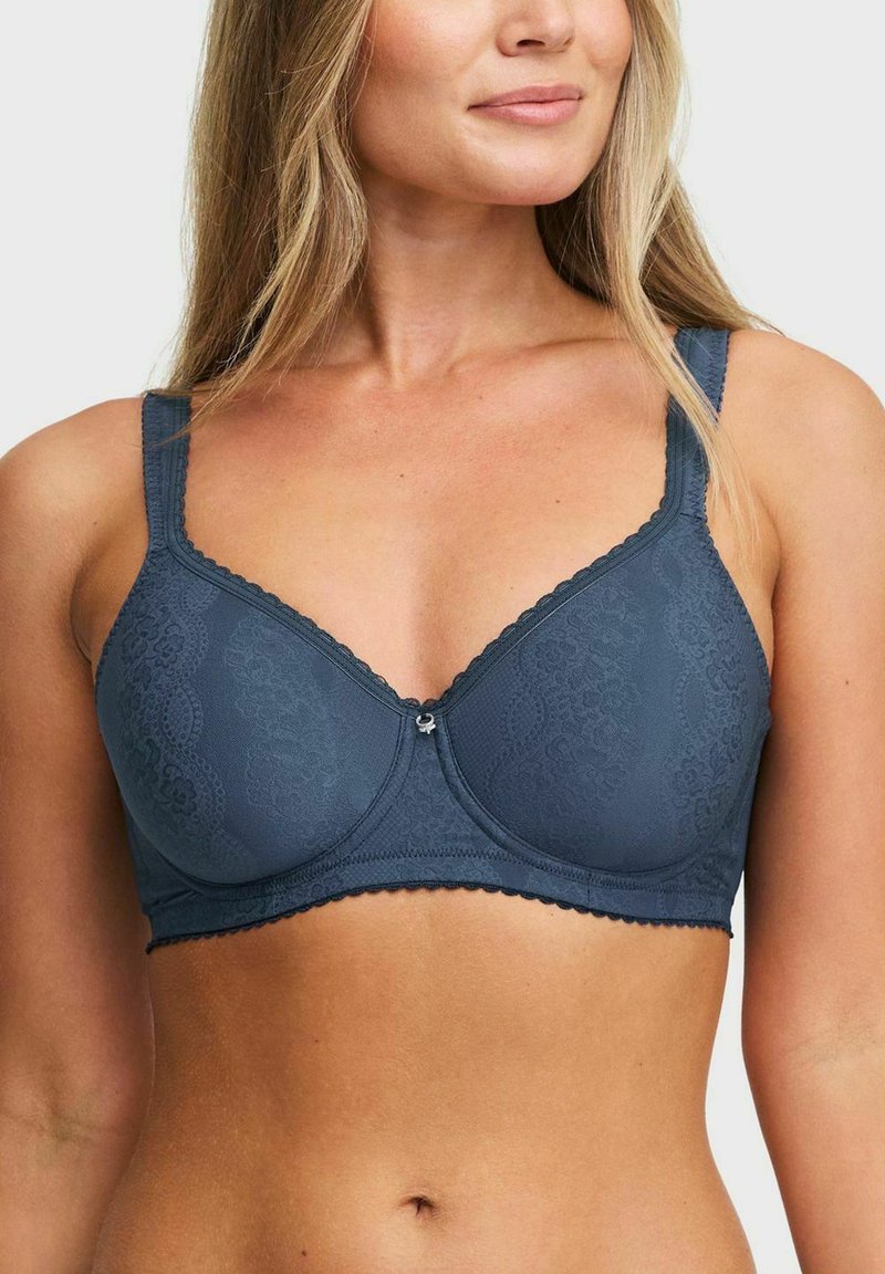 Vrouw in een blauwe kanten onderbh met verstelbare bandjes, zichtbaar van midden borst tot midden taille, tegen een effen achtergrond.