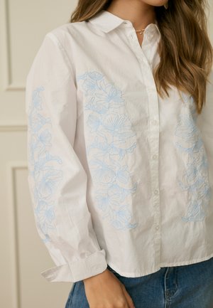 Femme portant une chemise blanche à boutons avec une broderie florale bleu clair sur le devant et les manches, associée à un jean en denim bleu.