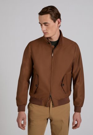 Uomo con indosso una giacca marrone con cerniera, tasche con patta e bottoni, polsini a costine, abbinata a pantaloni marrone chiaro, che guarda verso il basso di lato.