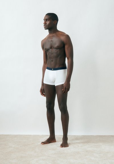 Μυώδης άνδρας που στέκεται ξυπόλητος σε χαλί, φοράει λευκά ανδρικά εσώρουχα με σκούρο μέση, κοιτάζει προς τα αριστερά με απλό φωτεινό φόντο.