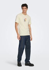 Μπλούζα από βαμβάκι σε κρεμ χρώμα με γραφικό λουλουδιών σε ροζ, κίτρινο, μπλε και μαύρο, τοποθετημένο στο κέντρο του στήθους, σε συνδυασμό με παντελόνι από σκούρο μπλε τζιν.