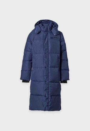 Jeune homme portant un long manteau matelassé bleu marine avec capuche, un sweat-shirt noir, un pantalon large sombre et des baskets noires, debout avec une main dans la poche.