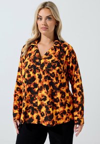 Donna che indossa una camicia a maniche lunghe con un motivo tie-dye arancione e nero, abbinata a pantaloni neri, in piedi contro uno sfondo bianco.