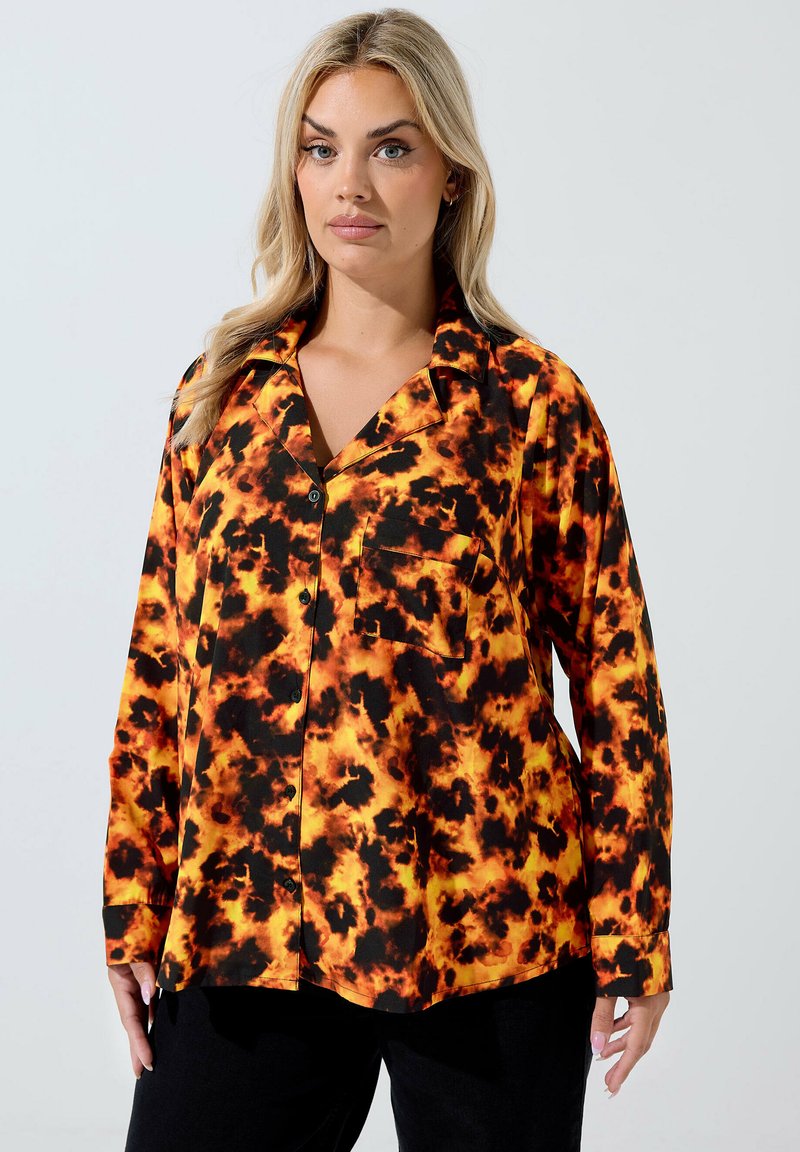 Donna che indossa una camicia a maniche lunghe con un motivo tie-dye arancione e nero, abbinata a pantaloni neri, in piedi contro uno sfondo bianco.