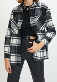 Camicia in flanella a quadri in bianco e nero con maniche lunghe, taschino sul petto e chiusura a bottoni, abbinata a pantaloni in ecopelle nera a vita alta.