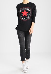 Person in einem schwarzen Converse All Star Langarmshirt, mit verwaschenen schwarzen Jeans mit kleinen Nieten und weißen High-Top Sneakern, der nach vorne geht.