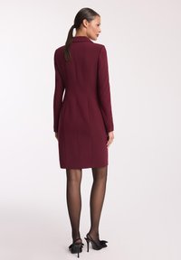 Burgundowa, dopasowana sukienka z długimi rękawami, z wyciętym kołnierzykiem, detalami wzdłuż szwu na plecach oraz guzikami na mankietach, zestawiona z czarnymi butami na obcasie typu slingback.