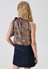 Top senza maniche in metallo color maroon con un laccio morbido sulla spalla, abbinato a una gonna in denim scuro con vita strutturata e tessuto testurizzato.