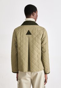 Olive green quilted jacket with a triangular accent on the back, paired with a cream-colored turtleneck. Smooth texture and structured design.
