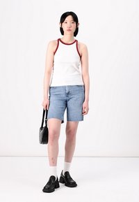 Balta juostuota tank top su bordo apvadu, šviesiai mėlyni džinsiniai šortai, juodi masyvūs loaferiai, balti kojinukai, laikant juodą krepšį. Vienalytis fonas.