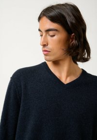 Donkerblauwe v-hals sweater van zacht materiaal, met een gebreid patroon en een ontspannen pasvorm, gepresenteerd door een model.