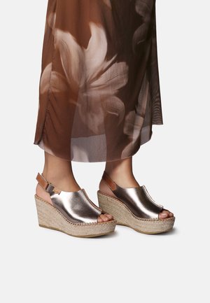 Ben iført metalliske kilehælssandaler med åbne tæer og ankelremme, kombineret med et semitransparent brunt blomsterprintet skørt.