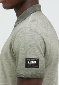 Kurzarm-Polohemd in Olivgrün mit strukturierter Oberfläche und einem dunkelgrünen Kragen. Verfügt über ein schwarzes Logopatch am Ärmel.