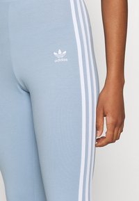 Detailní záběr osoby oblečené v světle modrých legínách Adidas s bílými třemi pruhy a logem, přičemž její pravá ruka je uvolněně položená podél těla.