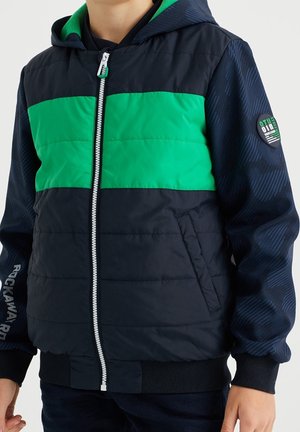 Chaqueta acolchada en azul marino con una franja horizontal verde. Cuenta con capucha, cierre de cremallera, puños elásticos y un parche en la manga.