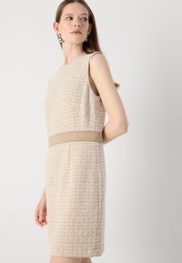Beige, teksturoitu, hihaton mekko, jossa on istuva siluetti ja leveä beige vyö vyötäröllä. Yksinkertainen, siisti muotoilu, jossa on vähäiset yksityiskohdat.