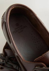 Mocassin en cuir marron avec semelle intérieure perforée estampillée "Made in Portugal Next" et détail décoratif en lacet sur un fond clair.