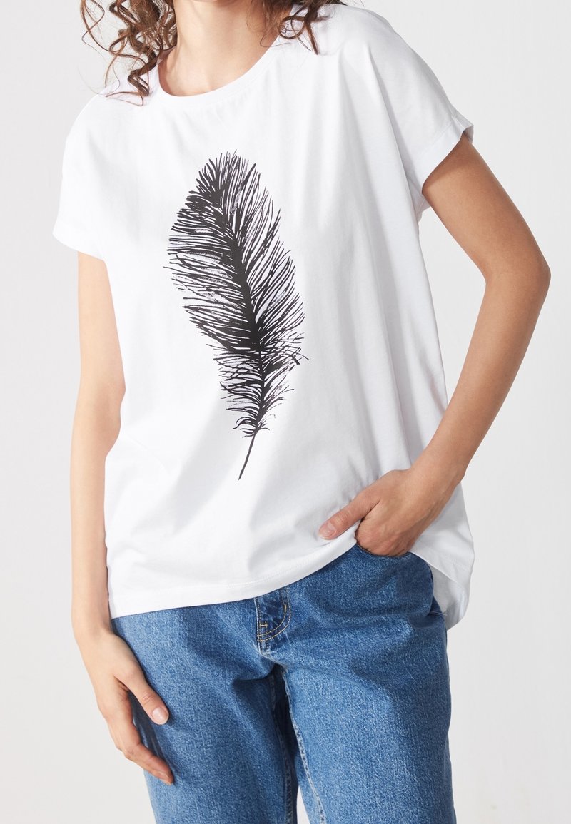 Femme portant un t-shirt blanc avec un grand motif plume noire, associée à un jean bleu, debout avec une main dans la poche.