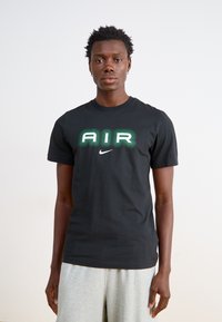Juodas medvilnės marškinėliai su žaliu 'AIR' logotipu ir mažu baltu Nike swoosh centruojant. Modelis dėvi šviesiai pilkas šortus.