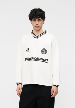 Junger Mann mit lockigem dunklem Haar trägt ein weißes langärmeliges Hemd mit schwarzem gestreiftem Kragen und dem Text "playa blanca" auf der Vorderseite, schwarze Hose.