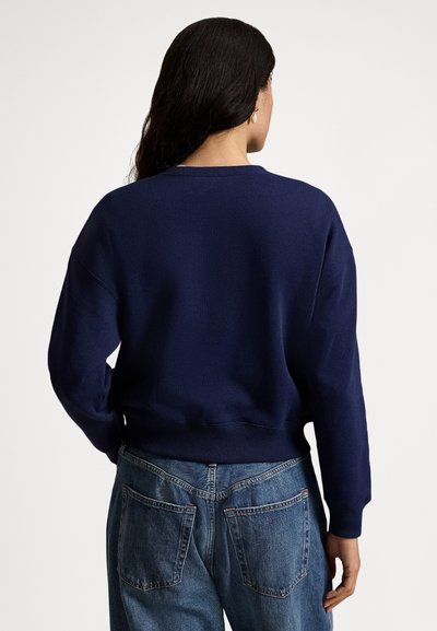 Vrouw in een marineblauwe trui met lange mouwen en een blauwe spijkerbroek, gezien vanaf de achterkant tegen een effen witte achtergrond.