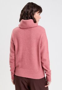 Pull en col roulé rose, à côtes, avec une coupe décontractée, des manches longues et une finition texturée, associé à un pantalon marron foncé.