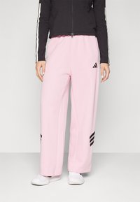 Lys rosa joggebukser med avslappet passform, med svarte Adidas-striper på øvre lår og en svart zip-up jakke med hvite striper.