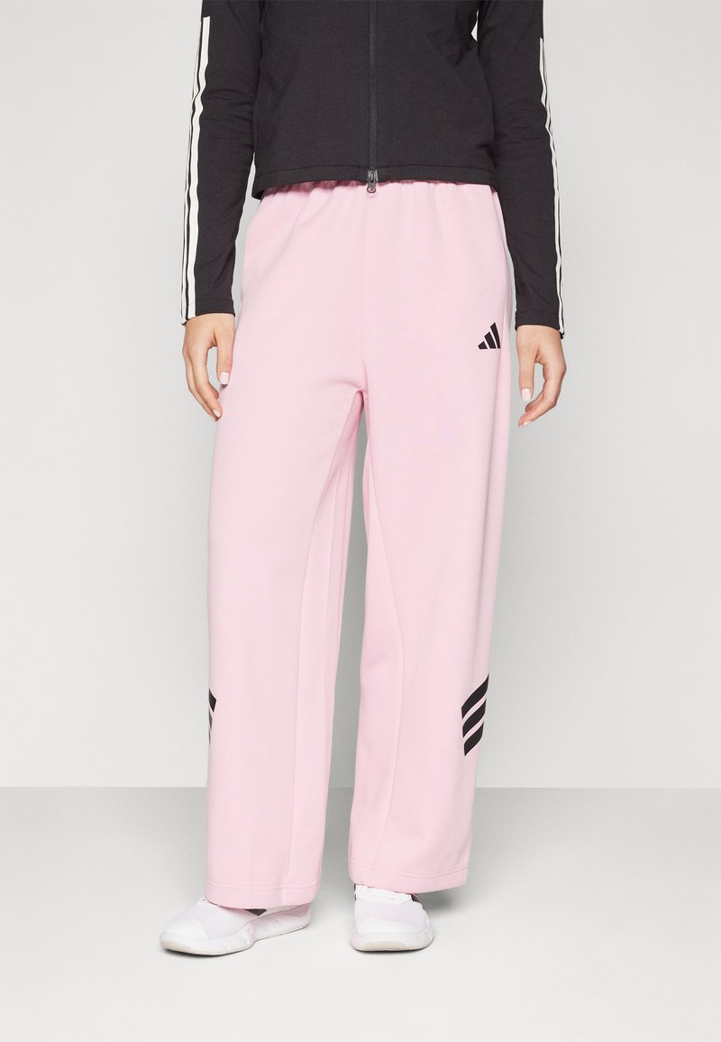 Lys rosa joggebukser med avslappet passform, med svarte Adidas-striper på øvre lår og en svart zip-up jakke med hvite striper.