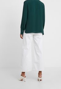 Blusa di seta verde con maniche lunghe e polsini abbottonati, abbinata a pantaloni bianchi a gamba larga e mule bianchi con tacco. Tessuto dalla consistenza liscia.