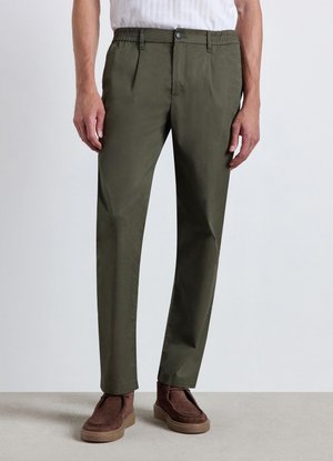 Uomo che indossa pantaloni dritti verde oliva con vita elastica e scarpe scamosciate marroni, in piedi su un pavimento neutro.