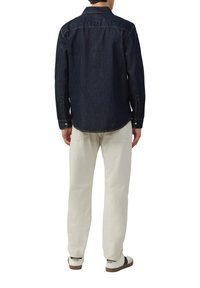 Donkerdenim overhemd met lange mouwen, puntige kraag en zijnaaden. Gecombineerd met lichtgekleurde broek en casual schoenen.
