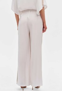 Lichtbeige wijde broek gemaakt van glad, vloeiend materiaal. Voorzien van een elastische tailleband en zijsplitten nabij de zoom voor extra bewegingsvrijheid.
