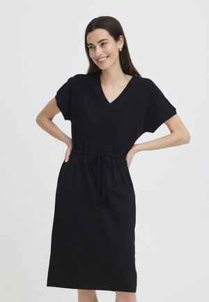 Mujer sonriendo, vestida con un vestido negro de largo medio, mangas cortas y cintura con cordón, con las manos apoyadas en las caderas.