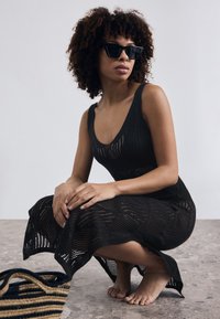 Abito in maglia nera con scollatura profonda e motivo intricato, abbinato a occhiali da sole neri angolari. Una borsa intrecciata con strisce nere e beige nelle vicinanze.