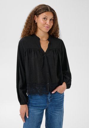 Femme aux cheveux bouclés portant un chemisier noir à manches longues avec bordure en dentelle et un jean bleu, posant avec une main dans la poche sur fond blanc.