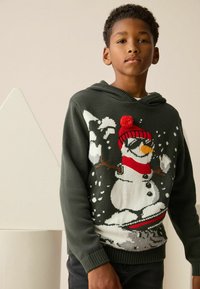 Grüne Strickjacke mit einem Schneemann-Design, rotem Hut mit Pompon und Schneedetails, kombiniert mit dunklen Jeans.