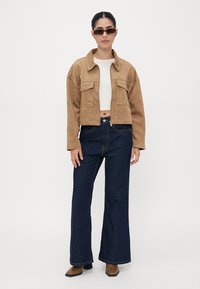 Kort brun mockajacka med två bröstfickor, tillsammans med mörkblå utsvängda jeans och bruna klackade ankelstövlar.