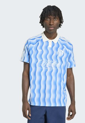 Junger Mann mit dunkler Haut und Dreadlocks, der ein blau-weiß gestreiftes Adidas-Polo mit wellenförmigem Muster und dunkelblaue Hosen trägt, steht frontal.