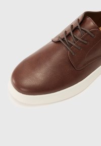 Chaussure en cuir marron avec une surface texturée, bout arrondi et lacets marron. Semelle en caoutchouc blanche avec des détails de couture.