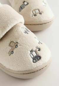 Chaussons en velours beige avec des véhicules de construction brodés, y compris un camion-benne et une pelleteuse sur la partie supérieure avant.