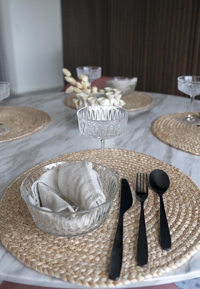 Eine klare Glasschüssel mit gefalteten Stoffservietten, schwarzem Besteck und einem strukturierten, gewobenen Platzdeckchen auf einer Marmortheke. Elegantes Glasgeschirr im Hintergrund.