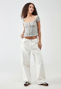 Kvinne med krøllete hår iført en grå kortermet crop-top, vide hvite bukser og svarte og beige sneakers, stående mot en ensfarget bakgrunn.