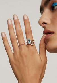 Tres anillos en una mano: uno presenta una gran piedra preciosa de color azul claro, otro tiene una piedra rectangular y un tercero es una banda delgada con pequeños acentos rojos.