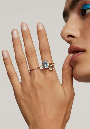 Tres anillos en una mano: uno presenta una gran piedra preciosa de color azul claro, otro tiene una piedra rectangular y un tercero es una banda delgada con pequeños acentos rojos.