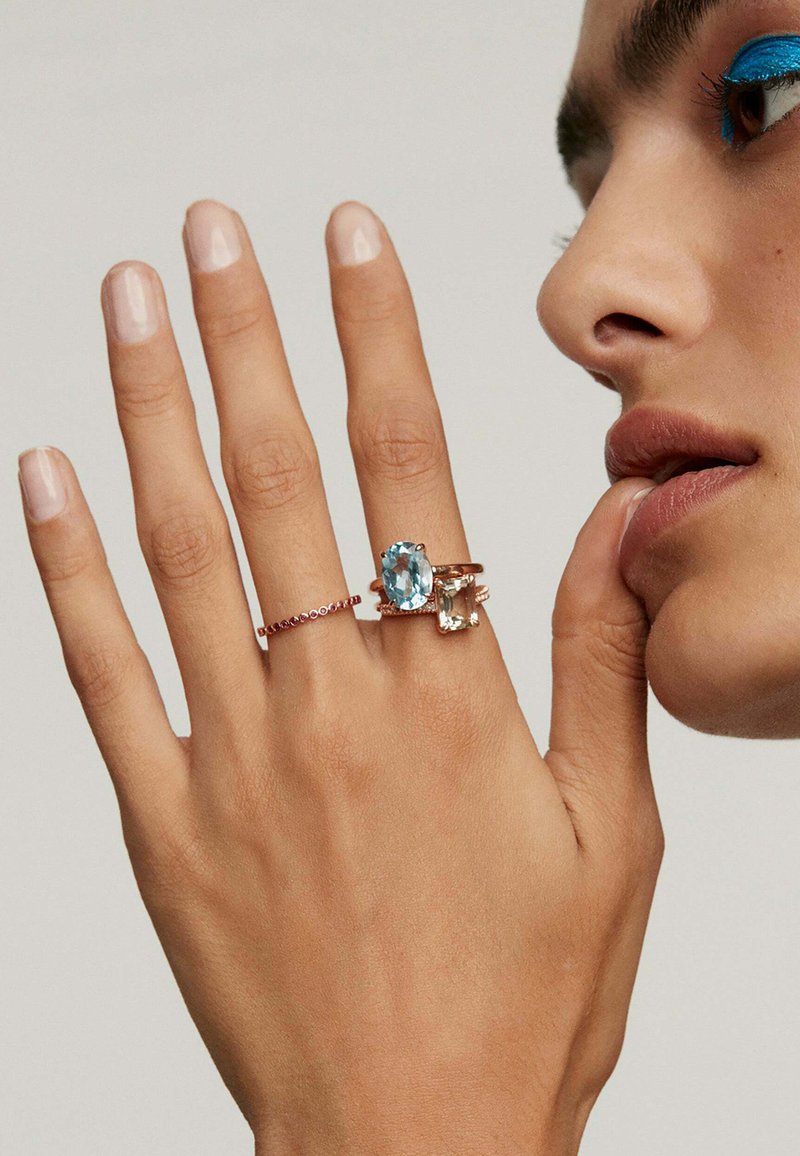 Tres anillos en una mano: uno presenta una gran piedra preciosa de color azul claro, otro tiene una piedra rectangular y un tercero es una banda delgada con pequeños acentos rojos.