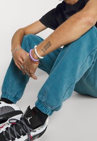 Tyrkysové tepláky s elastickými manžetami, doplněné o černobílé tenisky s texturovaným designem. Na zápěstí několik barevných korálkových náramků.